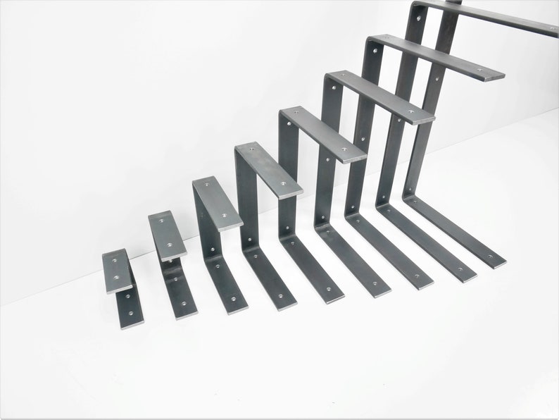K&ouml;nnte beinhalten: Ein Satz von neun Metall-Regalb&ouml;cken in verschiedenen Gr&ouml;&szlig;en. Die B&ouml;cke sind aus Stahl und haben eine rohe, unfertige Optik. Sie sind in einem versetzten Muster angeordnet, wobei der gr&ouml;&szlig;te Bock oben und der kleinste Bock unten ist.