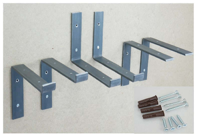 Puede incluir: Cinco soportes de estante de metal plateado con bordes redondeados y agujeros para tornillos. Los soportes est&aacute;n dispuestos en un patr&oacute;n escalonado sobre un fondo gris. Un juego de tacos de pl&aacute;stico marr&oacute;n y tornillos plateados se muestra en la esquina inferior derecha de la imagen.