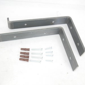 Puede incluir: Dos soportes de estante de metal con cuatro tornillos de montaje y cuatro tacos de pl&aacute;stico. Los soportes son de color gris oscuro y tienen una forma ligeramente curvada.