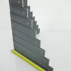 May include: A stack of gray metal L-brackets with pre-drilled holes, arranged in a stepped pattern. A yellow measuring tape is placed along the bottom, showing measurements in inches. The brackets are made of a sturdy metal.
