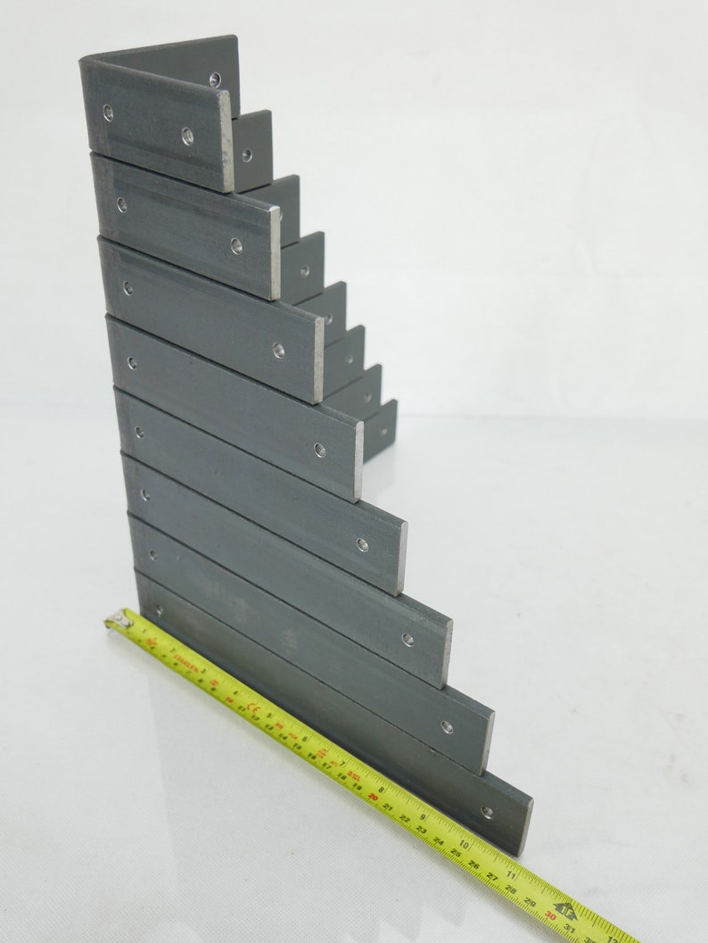 May include: A stack of gray metal L-brackets with pre-drilled holes, arranged in a stepped pattern. A yellow measuring tape is placed along the bottom, showing measurements in inches. The brackets are made of a sturdy metal.