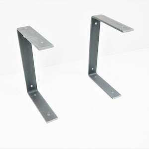 K&ouml;nnte beinhalten: Zwei silberne Metall-Regalhalterungen, jede mit einem 90-Grad-Winkel und einer flachen, rechteckigen Basis.