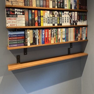 May include: Wooden bookshelves with multiple rows of books. The books have various colors and titles, including authors like Jo Nesbo and Dennis Lehane. The shelves are supported by dark metal brackets, mounted on a gray wall. The shelves are made of light-colored wood.