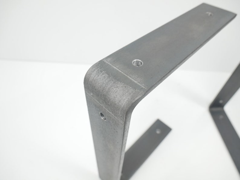 K&ouml;nnte beinhalten: Ein Paar Tischbeine aus Metall mit grauer Oberfl&auml;che. Die Beine sind L-f&ouml;rmig und haben eine abgerundete Ecke. In jedes Bein sind drei L&ouml;cher gebohrt.