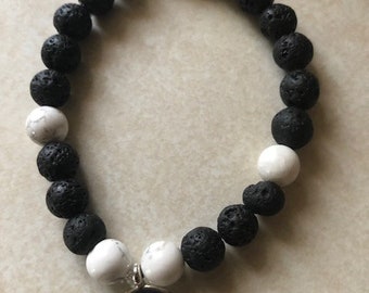 May include: A black and white beaded bracelet with a silver charm featuring a meditating figure. The bracelet is made with lava rock and white beads.