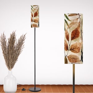 May include: A floor lamp with a black metal stand and a cylindrical lampshade. The lampshade features a pattern of brown and green leaves on a cream background. A white vase with dried plants is on the left.