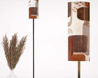 May include: A floor lamp with a black metal frame and a cylindrical lampshade. The lampshade features a geometric pattern in shades of brown, beige, and rust. The lamp is positioned next to a white vase with dried ornamental grass.