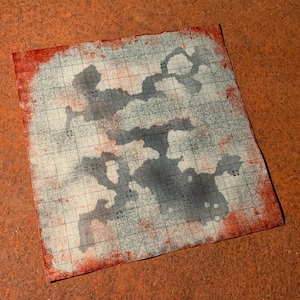 Puede incluir: Un mapa cuadrado, blanquecino, con un patrón de cuadrícula y manchas de color gris oscuro, posiblemente representando una mazmorra o terreno. Los bordes están manchados de rojo, y el mapa está sobre una superficie texturizada, marrón anaranjado.