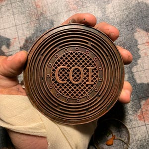 Puede incluir: Un objeto metálico redondo de color bronce con las letras "COI" en el centro. El objeto tiene crestas circulares concéntricas y pequeños remaches circulares en el borde. Se sostiene en una mano con una venda.