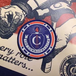 May include: A blue and white embroidered patch with a red border. The patch features a stylised letter "C" with a torch above it and the text "The Commonwealth" around it.