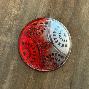 May include: A circular shield featuring a split design with a vibrant red side and a metallic silver side. The surface is decorated with black geometric fan-shaped patterns and small circular rivets along the outer edge of the round shield.