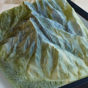 May include: A 3D model of a mountain range, with a textured surface that appears to be made of plaster or clay. The mountain range is painted in shades of green, brown, and white, giving it a realistic appearance.