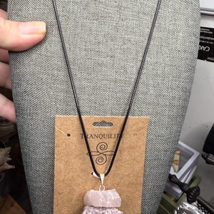 Peut inclure: Un collier pendentif en cristal superposé avec un cordon noir. Le pendentif présente trois pierres de cristal brutes empilées dans des tons de rose, de lavande et de gris. Le collier est suspendu à une carte d'affichage avec le mot "TRANQUILITY" imprimé.