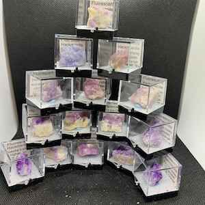 May include: A collection of small, clear plastic boxes containing various purple and pink mineral specimens. Each box has a label identifying the mineral and its origin, such as "Hackmanite, Valley, Afghanistan".