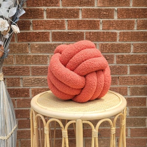 May include: A coral-colored knot pillow made of a soft, textured material. The decorative pillow is displayed on a small, round, light-colored wicker table. A tall vase with dried flowers is to the left, and a brick wall is in the background.