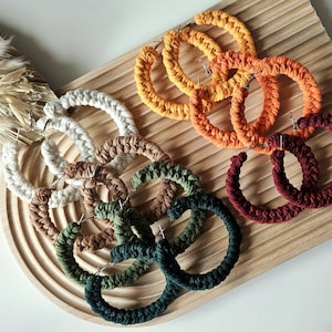 May include: A set of six pairs of crocheted hoop earrings in various colors, including white, brown, green, orange, and red. The earrings are displayed on a wooden surface.