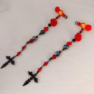 May include: Two black metal sword-shaped charms with red rose and black bead accents on a chain.