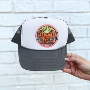 May include: A grey and white trucker hat with a circular patch featuring a sunset, mountains, cacti, and a bicycle. The patch has a pink, orange, and yellow colour scheme.