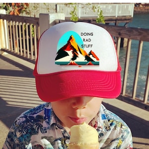 May include: A child wearing a red and white trucker hat with the text "Doing Rad Stuff" and a mountain graphic. The child is holding a yellow ice cream cone.
