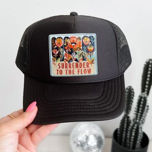 Peut inclure: Une casquette de type trucker noire avec un écusson rectangulaire sur le panneau avant. L'écusson présente un motif floral brodé coloré au-dessus du texte "SURRENDER TO THE FLOW" en police grasse. La casquette possède un filet et une visière.