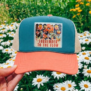 May include: A teal and coral trucker hat featuring a rectangular patch with an embroidered floral design and the text "SURRENDER TO THE FLOW" in red lettering. The hat has a mesh back panel and a structured front panel with a dark grey cord trim.