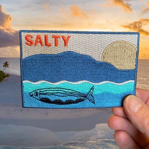 May include: Embroidered patch with the word "SALTY" in orange letters above a blue ocean scene with a fish. The patch features a blue mountain range, a tan sun, and a fish with black and blue details.