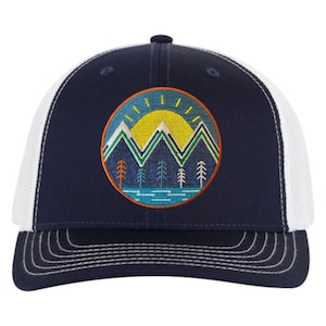 May include: A navy blue and white trucker hat with a circular embroidered patch featuring a mountain range, a sun, and trees. The patch has a blue, green, yellow, and orange color scheme.