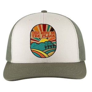 May include: A white and green trucker hat with a brown mesh back. The front of the hat features an embroidered patch with a retro-style sun, hills, and trees. The patch has the text "This must be the place" in orange and yellow.