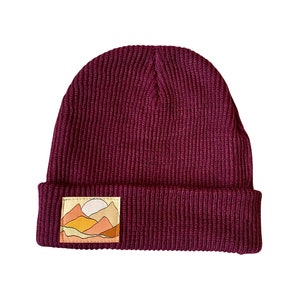 Può includere: Un berretto di lana color borgogna con una toppa che raffigura un paesaggio di tramonto con montagne e un sole.