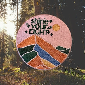 May include: A pink embroidered patch with a mountain range design and the text "shine your light" in green. The patch has a yellow sun and white stars.