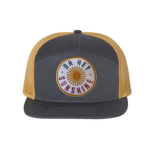 May include: A grey and gold trucker hat with a white circle patch that says "Oh Hey Sunshine" in colourful lettering. The patch features a yellow sun with red, green, and blue accents.