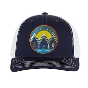 Puede incluir: Gorra de camionero azul marino con parte trasera de malla blanca y visera curva. La parte delantera presenta un diseño bordado circular de montañas, sol y árboles en tonos amarillo, verde, azul y naranja. Cierre de broche.