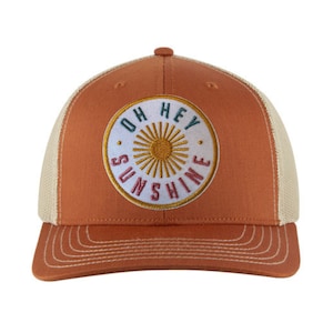 May include: A brown and beige trucker hat with a white circle patch that says "Oh Hey Sunshine" in colorful embroidery. The sun is depicted in the center of the circle.
