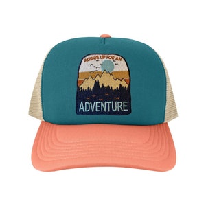 May include: A teal and peach trucker hat with a mesh back. The front panel has an embroidered patch with a mountain scene and the words "ALWAYS UP FOR AN ADVENTURE". The brim is peach-coloured.