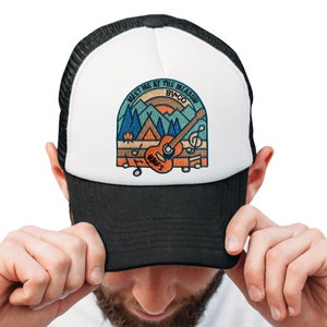 May include: A black and white trucker hat with a colourful embroidered patch. The patch features a guitar, musical notes, and the text "MEET ME AT THE MEADOW BV+CO". The design includes a mountain scene and a campfire.