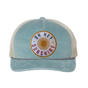 May include: A light blue corduroy trucker hat with a white mesh back. The hat features a circular patch with the words "OH HEY SUNSHINE" surrounding a gold sunburst design. A grey rope detail accents the brim.