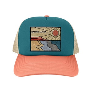 Peut inclure: Un chapeau de camionneur turquoise et corail avec un dos en maille beige. Le devant du chapeau présente un patch brodé avec une scène de coucher de soleil et le texte "NATURE LOVER".
