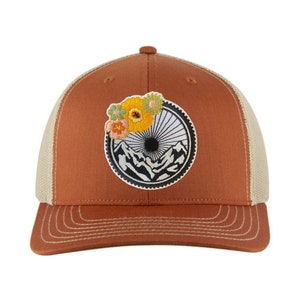 Puede incluir: Una gorra de camionero marrón y beige con un parche bordado en blanco y negro de una escena de montaña con un sol y flores.