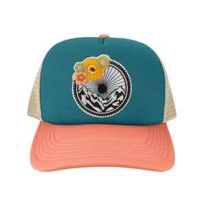 May include: Teal trucker hat with a peach-colored brim and a tan mesh back. The hat features an embroidered patch with a mountain scene, a bicycle wheel, and colorful flowers. The top button is peach-colored.