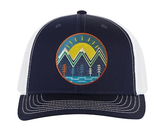 May include: A navy blue and white trucker hat with a circular embroidered patch featuring a mountain range, a sun, and trees. The patch has a blue, green, yellow, and orange color scheme.