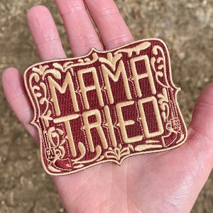 May include: A red embroidered patch with gold lettering that reads "Mama Tried". The patch has a decorative border with floral designs.