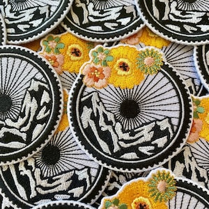 May include: A collection of embroidered patches featuring a black and white mountain scene with a sunburst design. Some patches have a white background and others have a yellow floral design with orange and green accents.