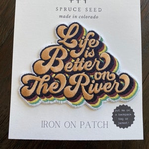May include: Iron-on patch with the text "Life is Better on the River" in a retro font with a rainbow color scheme. The patch is on a white card with the text "Spruce Seed made in Colorado" and "Iron on Patch".