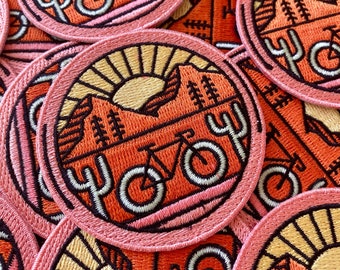 May include: Close-up of several embroidered patches featuring a sunset, mountains, cacti, and a bicycle. The patches are pink, orange, and yellow with black outlines. The text "Yoy" is embroidered on each patch.