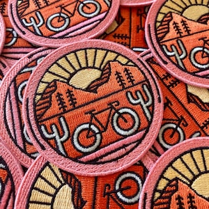 May include: Close-up of several embroidered patches featuring a sunset, mountains, cacti, and a bicycle. The patches are pink, orange, and yellow with black outlines. The text "Yoy" is embroidered on each patch.