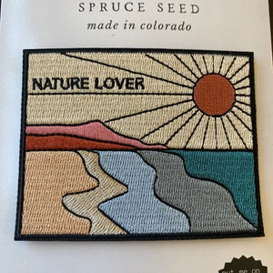 May include: Iron-on patch with a nature lover theme. It features a sunset over a beach with a mountain in the background. The text "NATURE LOVER" is embroidered in black on a beige background. The patch is surrounded by a black border.