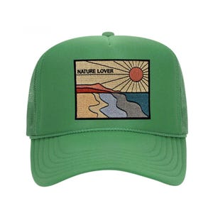 Puede incluir: Gorra de camionero verde con parte trasera de malla y visera curva. La parte delantera presenta un parche bordado con un diseño de paisaje, que incluye un sol, montañas y agua, con el texto "NATURE LOVER".