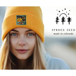 Pode incluir: Um gorro de malha amarelo com um remendo bordado colorido que representa uma cena de montanha com um sol, árvores e uma flor. O gorro é usado por uma pessoa com cabelo loiro. O texto "SPRUCE SEED made in colorado" é visível na imagem.