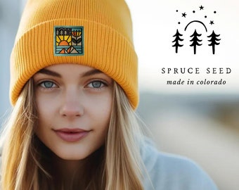 May include: A yellow knit beanie with a colorful embroidered patch depicting a mountain scene with a sun, trees, and a flower. The beanie is worn by a person with blonde hair. The text "SPRUCE SEED made in colorado" is visible in the image.