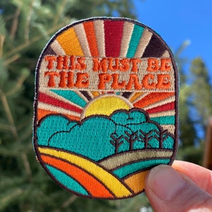 May include: A colorful embroidered patch with a retro sun, clouds, and trees. The text "This must be the place" is stitched in orange and brown.